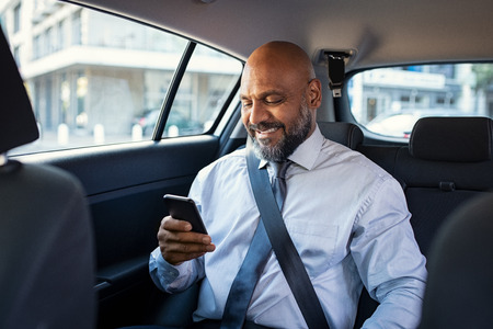 Successful african businessman working on phone smiling witting in car. Portrait of mature smiling business man in formal clothes using smartphone while sitting on back seat of luxury business car. Senior formal man reading confirmation mail on smartphone and smiling in a taxi.の写真素材