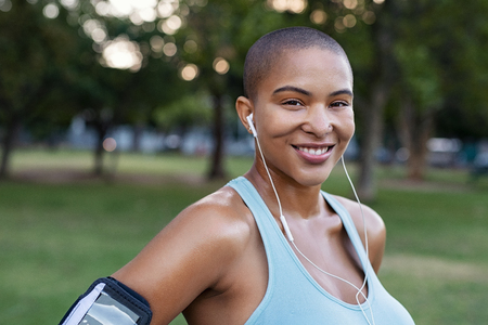 Portrait of a smiling curvy woman listening to music with earphones standing in park after running. Bald young woman looking at camera after fitness workout. Closeup face of satisfied sweating girl after sporty exercises.の写真素材