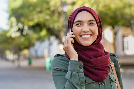 Beautiful and happy woman in hijab talking over mobile phone. Portrait of arabic woman in religious headscarf talking on cellphone. Smiling islamic girl with hijab looking away while talking over phone.の写真素材