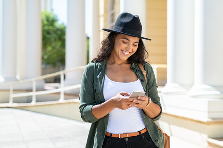 Cheerful young woman wearing black hat in city street typing a message. Hispanic trendy girl browsing internet on phone outdoor. Happy stylish girl using smartphone while travelling the city.の写真素材