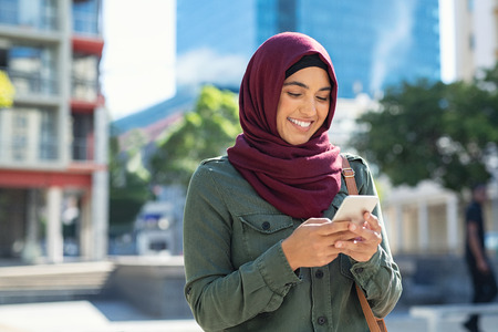 Pretty muslim woman using mobile phone outdoor. Arabic woman wearing hijab using smartphone on the street. Islamic girl texting a message phone in city centre.の写真素材
