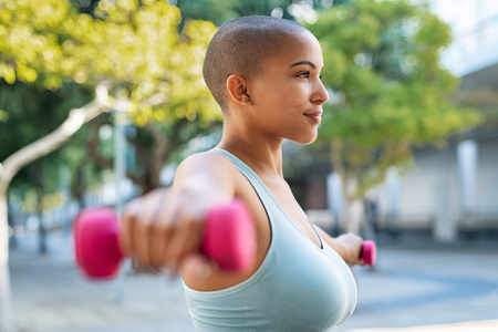 Active curvy woman using dumbbells in the park. Side view of young woman doing stretching exercise using dumbbells. Active athlete working out on fitness routine to lose weight.の写真素材