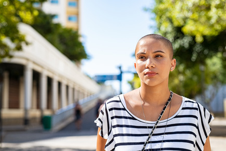 Serious young woman with bald hair lost in deep thoughts. Stylish woman in casual dress looking away thinking. Proud young girl suffering from cancer looking away while standing on road, copy space.の写真素材