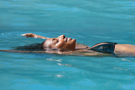 Young african american woman relaxing in swimming pool at spa resort. Portrait of beautiful black girl in bikini floating on water. Side view of cheerful tanned woman enjoying swimming at summer vacation.の写真素材