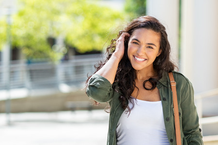 Happy young beautiful woman wearing green jacket and white shirt walking on the street. Portrait of cheerful university student looking at camera while adjusting curly hair with copy space. Latin stylish girl smiling while standing on street.の写真素材