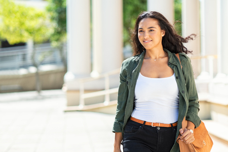 Young attractive woman walking in city street and looking away. Beautiful latin girl in casual with urban background. Hispanic casual woman smiling and walking on a bright sunny day with wind in the hair.の写真素材