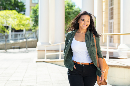 Pretty casual natural woman walking in urban street and looking away. Cheerful latin girl with bag travelling across the city. Beautiful young woman smiling outdoor.の写真素材