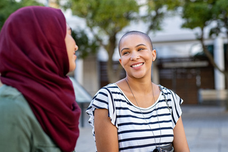 Cheerful african american woman in conversation with muslim girl. Happy multiethnic friends in street looking at each other while in a conversation. Beautiful woman with friend in hijab talking to each others.の写真素材