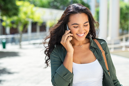 Cheerful young woman looking down on street while talking over smartphone. Smiling casual girl walking on street and talking on phone. Smiling businesswoman having pleasant conversation on mobile phone.の写真素材