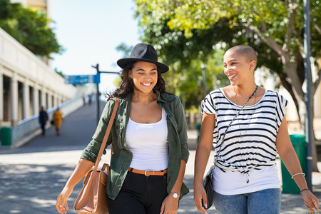 Young smiling women walking and talking while looking at each other outdoor. Happy best friends laughing and having fun while walking on city street. Two women friends sightseeing in summer while on vacation.の写真素材