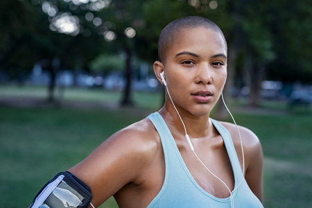 Portrait of motivated young woman resting after jogging. Beautiful focused bald girl take a break after run. Determined runner listening to music while jogging and looking at camera.の写真素材