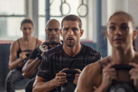 Portrait of fitness man lifting kettle bell with young women in fitness center. Active guy with group of people in gym doing weight lifting with kettlebell. Determined people in a row squatting in cross fit gym.の写真素材