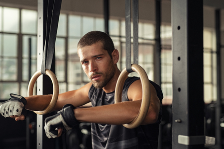 Tired handsome strong man holding on to gymnastic rings at cross fit gym. Closeup portrait of fitness man taking a break after intense workout at gym. Young man relaxing with arms in gymnastic rings.の写真素材