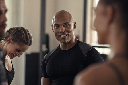 Friends in sportswear talking and laughing together in gym after a workout. Group of four multiethnic people relaxing in conversation after crossfit training. Black sweaty guy and young women talking to each other while resting after intensive training in cross fit gym.の写真素材