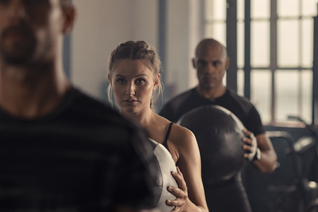 Determined young woman doing warmup exercise with her class in gym. Fit man and woman doing weight lifting in fitness center using medicine ball. Portrait of beautiful woman doing squat exercise with heavy ball.の写真素材
