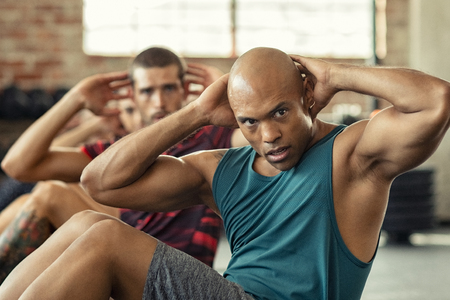 Muscular african american man doing sit ups at gym with other people in background. Mature black man doing abs workout with class at crossfit gym. Muscular guy doing crunches.の写真素材