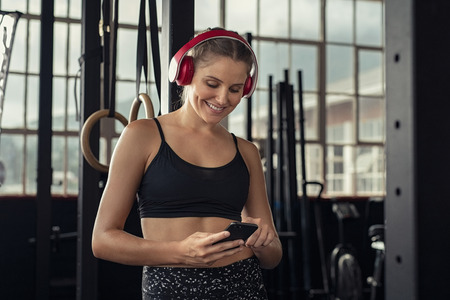 Young woman with smartphone listening music at gym with wifi headset. Smiling beautiful fit girl relaxing after exercise. Happy sporty woman with cellphone and cableless connection of headphones take a break from workout.の写真素材