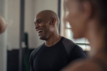 Bald mature man enjoying a break while chatting at gym. Closeup of african american athlete laughing while in conversation with woman in fitness center. Carefree black trainer in sportswear resting after workout exercise with class.の写真素材