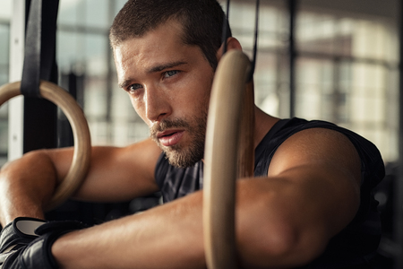 Closeup of young man resting after gymnastic ring exercise at crossfit gym. Fitness guy thinking while leaning at dip rings and looking away. Determined man relaxing after workout training.の写真素材