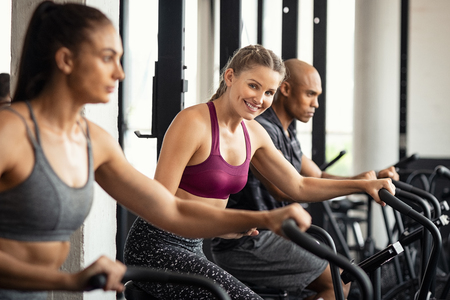 Fit woman working out with class on stationary bike at gym. Portrait of young beautiful woman smiling and looking at camera while cycling at gym. Happy cheerful athletes in a row training on exercise bike.の写真素材