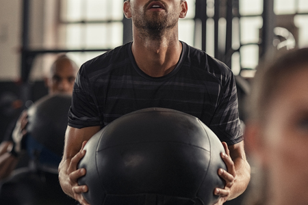Closeup of sportsman doing weight lifting with heavy medicine ball. Determined man doing crossfit training in fitness center. Young man doing intensive squat workout at gym with fitness ball.の写真素材