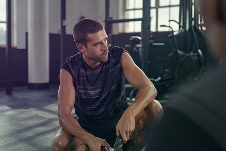 Tired young sportsman resting after training with coach. Handsome man sitting with battle ropes relaxing after exercise in crossfit gym. Exhaustive guy taking a break while looking away at industrial gym.の写真素材