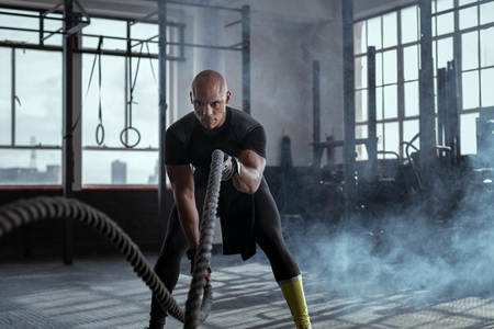 Bald athlete doing battle rope exercise at crossfit gym. Concentrated african man doing cross fit exercise while working out in gym. Focused man in sportswear training exercise.の写真素材