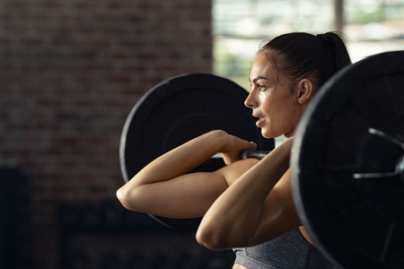 Fitness woman doing shoulder press exercise with a weight bar at crossfit gym. Muscular woman in gym doing heavy weight exercises. Concentrated athlete doing barbell lifting at health club with copy space.の写真素材