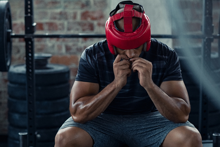 African american man in red boxing head gear putting gumshield. Determined boxer in sportswear sitting near boxing ring. Young man preparing for boxe practice with protective helmet.の写真素材
