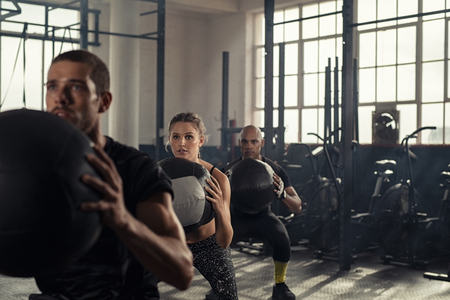 Group of three people doing squats while holding heavy medecine balls. Young men and fitness woman doing bodyweight exercises using weight ball. People in a row squatting with weighted balls at crossfit gym.の写真素材