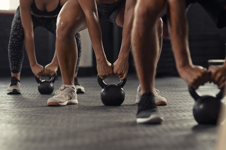 Closeup of young man and fit woman hands lifting kettle bell while squatting at gym. Athlete people in fitness center doing weight lifting with kettlebell. Group of three young athlete doing crossfit training.の写真素材