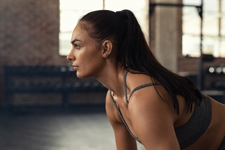 Young woman doing squat exercise while looking away at gym. Closeup of muscular girl while exercising in fitness center. Fit and concentrated woman doing crossfit workout.の写真素材