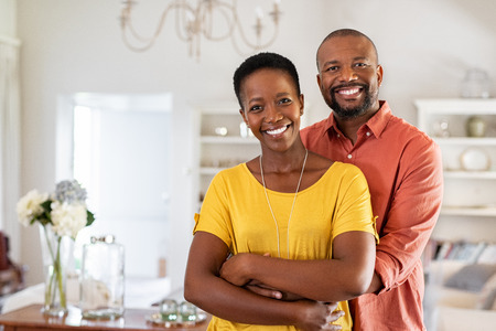Smiling mature couple holding each other at home. Loving african couple standing in living room embracing and looking at camera. Husband hugging wife from stomach at new apartment with copy space.の写真素材