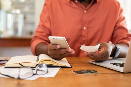 Closeup of african man hands using phone to calculate expenses. Man checking invoice balance on mobile phone app. Close up hands of black guy taking a closer look at his budget and calculating credit card bills.の写真素材