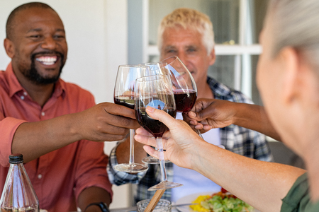 Multiethnic friends cheering with wine glasses under the patio during lunch. Happy mature and senior people having fun together while raising a toast with glasses of red wine. Generation couple toasting with red wine at picnic table to celebrate.の写真素材