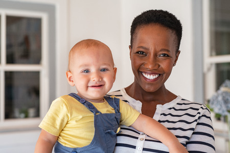 Portrait of african mature nanny with baby boy looking at camera. Smiling black mother holding adopted child and smiling together. Portrait of woman relaxing at home with son.