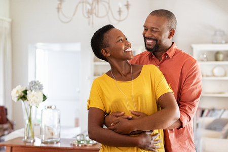 Mature married couple embracing in living room while looking at each other. Newly wed african couple in love hugging at home. Romantic husband with beautiful wife in love at home with copy space.の写真素材