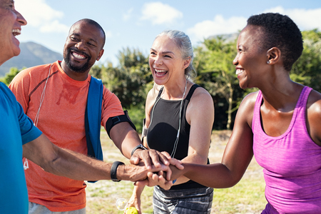 Laughing senior and multiethnic sports people putting hands together at park. Happy group of men and women smiling and stacking hands outdoor. Multiethnic sweaty team cheering after intense training.の写真素材