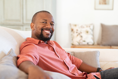 Portrait of happy mature african american man sitting on couch. Satisfied black man looking at camera while resting in living room. Handsome senior relaxing at home on the sofa.の写真素材