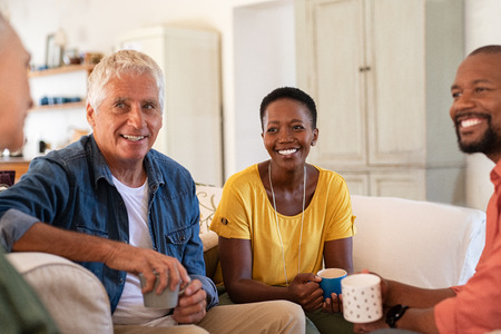 Group of multiethnic friends talking and drinking tea at home. Mature and senior couple chatting together while sitting on couch. Happy man and smiling woman looking at each other while in conversation at home and drinking coffee.の写真素材