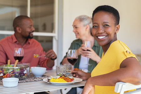 Happy mature african woman looking at camera while having lunch with her friends in background. Portrait of laughing black woman enjoying brunch with senior people. Smiling casual lady and multiethnic couple eating and drinking together.の写真素材