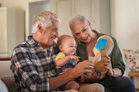 Toddler playing with old grandparents and teddy bear stuff toy. Cheerful grandfather and smiling grandmother showing soft toy to grandchild. Cute toddler boy playing with senior man and woman sitting on couch.の写真素材