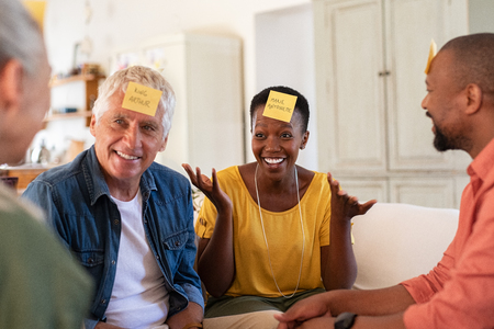 Group of multiethnic mature friends playing guess sticky head game at home. Men and smiling women enjoying charades game session in living room. African woman trying to make her friend guess the character.の写真素材