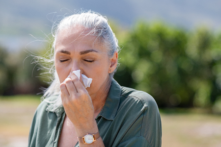 Senior woman with tissue having flu or allergy at park. Mature sick woman with grey hair suffering from cold outdoor. Old ill lady with closed eyes blowing nose with copy space.の写真素材