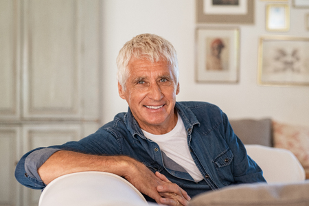 Happy senior man sitting on comfortable sofa in living room. Old man with grey hair relaxing on couch and smiling while looking at camera. Elderly man enjoying retirement at home.の写真素材