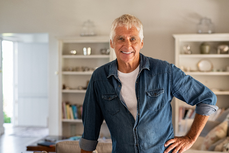 Happy senior man standing in living room and looking at camera. Portrait of cheerful old man with blue denim shirt at home relaxing. Satisfied man enjoying the retirement at home.の写真素材