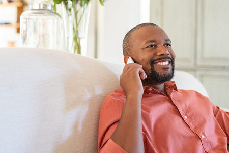 Cheerful mature man talking on smartphone. Black man at home relaxing while talking on cellphone. Happy african american guy sitting on couch in conversation through mobile phone with copy space.の写真素材