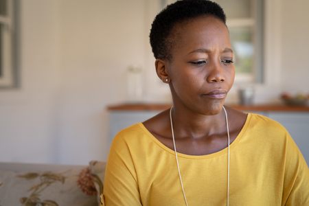 Depressed african woman sitting on couch. Mature lonely woman sitting at home and looking down. Depressed lady thinking. Solitude concept.の写真素材