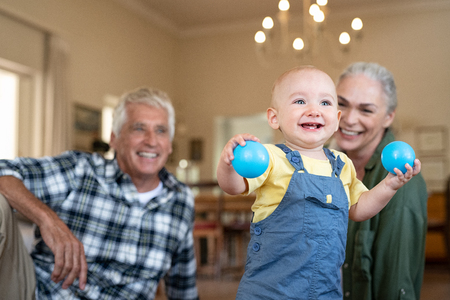 Happy baby playing with blue balls on floor at home with grandparents in background. Cute boy standing and holding toy while looking up. Excited toddler enjoying playing with senior man and old woman at home. Little boy standing and walking.の写真素材