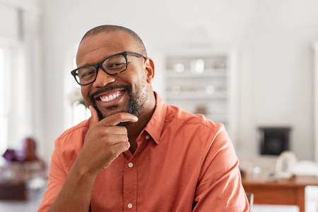 Cheerful black mature man wearing spectacles looking at camera. African businessman with hand on chin smiling while sitting at home. Happy man with eyeglasses enjoying new project.の写真素材